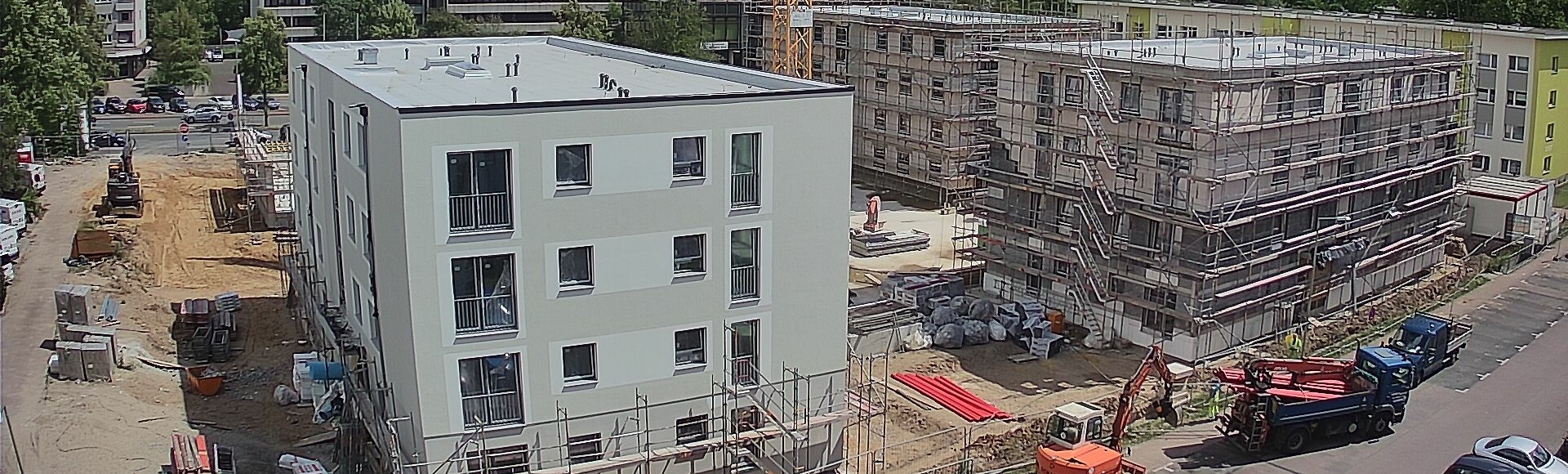 Baustelle mit Kran und im Bau befindlichen Gebäuden an einem sonnigen Tag, umgeben von Bäumen und Wohnanlagen.