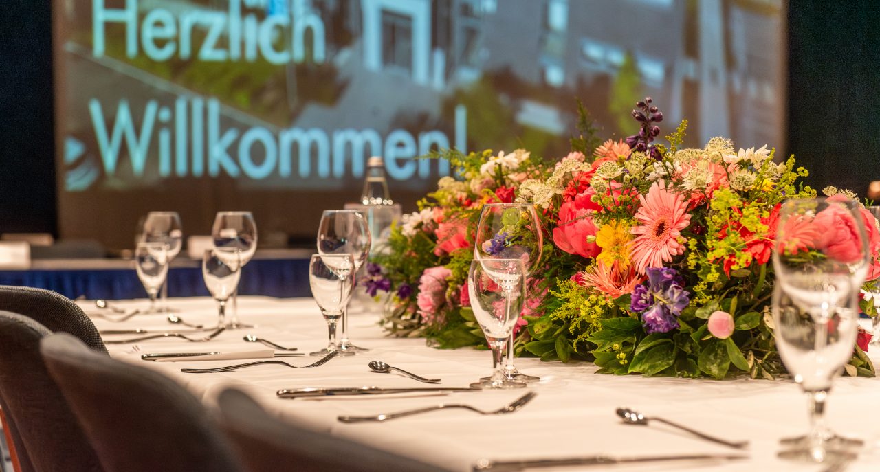 Elegant gedeckter Tisch mit Blumengesteck in der Mitte, umgeben von Weingläsern und Besteck; im Hintergrund unscharf ein Bildschirm mit der Begrüßung „Herzlich Willkommen“.
