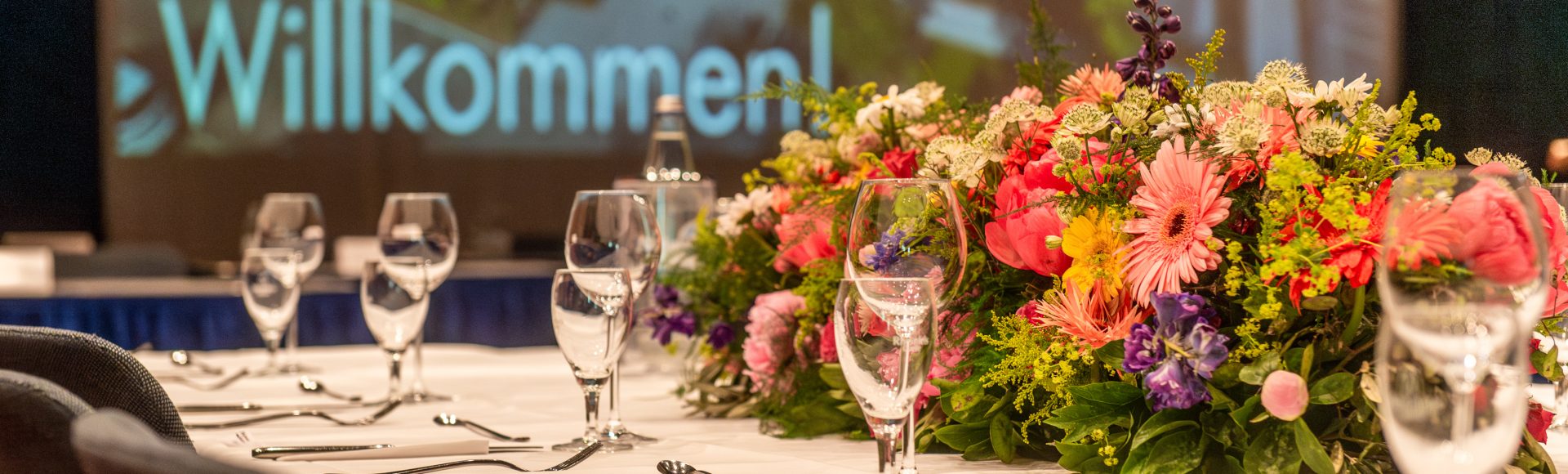 Elegant gedeckter Tisch mit Blumengesteck in der Mitte, umgeben von Weingläsern und Besteck; im Hintergrund unscharf ein Bildschirm mit der Begrüßung „Herzlich Willkommen“.